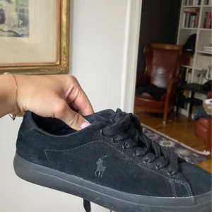 Svarta sneakers från Polo Ralph Lauren - Svarta sneakers från Polo Ralph Lauren i mocka med klassisk låg profil och rund tå. Skorna har svart snörning, diskret logga på sidan och bekväm platt sula. Perfekta för en clean och stilren look. Storlek 37-38