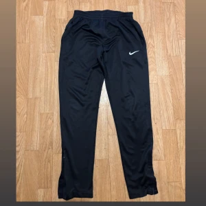 Svarta träningsbyxor från Nike - Svarta träningsbyxor från Nike med klassisk vit swoosh-logga på låret. Byxorna har elastisk midja, raka ben och är tillverkade i ett glansigt, lätt polyestermaterial. Perfekta för träning eller chill, med fickor på sidorna och diskret logga bak.