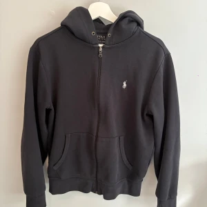 Svart hoodie från Polo Ralph Lauren - Svart hoodie från Polo Ralph Lauren med dragkedja framtill och klassisk huva. Tröjan har långa ärmar, känguruficka och den ikoniska broderade loggan på bröstet. Perfekt för en chill och stilren look. Pris kan diskuteras vid snabb affär