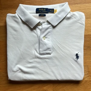 Ralph Lauren Pikétröja – Vit – Strl M (Slim Fit) – Klassisk & Tidlös - Lyft din stil med denna ikoniska Ralph Lauren-piké. Tillverkad i mjuk bomull med en smickrande slim fit – perfekt för både vardag och mer uppklädda tillfällen. 	•	Märke: Ralph Lauren 	•	Färg: Vit 	•	Storlek: M (Slim Fit) 	•	Mått: Bröstvidd 47 cm, Längd 65 cm 	•	Skick: Utmärkt – inga fläckar, hål eller slitage.  En tidlös klassiker och ett måste i en välklädd garderob.
