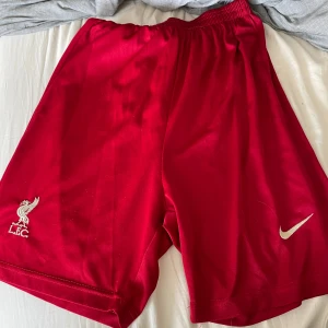 Röda Nike Liverpool FC shorts - Snygga röda shorts från Nike med Liverpool FC-logga på ena benet och klassisk Nike-swoosh på det andra. De har elastisk midja och är gjorda i ett lätt, sportigt material som passar perfekt för fotboll eller träning.
