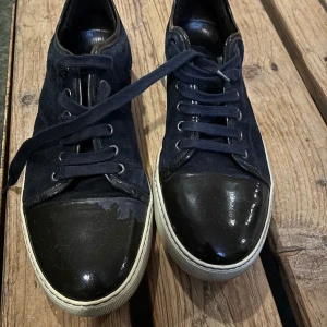 Svarta sneakers med lackad tå - Snygga svarta sneakers med ovandel i mocka och tåparti i blankt lack. Skorna har vita sulor och svarta skosnören. Klassisk rund tå och stilren design som passar till det mesta. Ena skon är lite skadad fram på högra tån