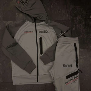 Grå hoodie från Hoodrich med dragkedja - Snygg grå hoodie från Hoodrich med svarta detaljer och tryck på bröstet. Tröjan har huva med snörning, hel dragkedja och en extra ficka med dragkedja på framsidan. Perfekt för en streetwear-look.