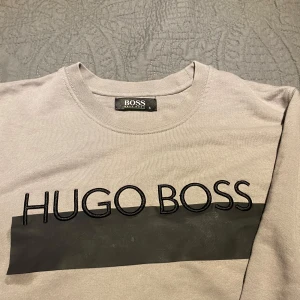 Beige sweatshirt från Hugo Boss - Snygg beige sweatshirt från Hugo Boss med svart logga broderad över bröstet och svart band. Tröjan har rund halsringning, långa ärmar och ribbade muddar. Perfekt för en clean och stilren look. Materialet är mjuk bomull som känns skönt mot huden.