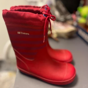 Röda randiga stövlar från Tretorn - Säljer ett par röda stövlar från Tretorn med snygga horisontella ränder och dragsko upptill. Skorna har rund tå och är tillverkade i ett tåligt syntetmaterial med textil upptill. Perfekta för regniga dagar och utomhusaktiviteter.