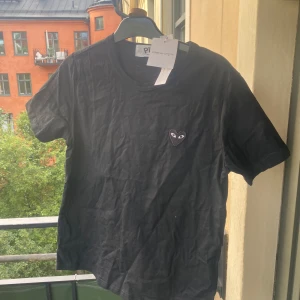 Svart Comme des Garçons Play t-shirt - Svart t-shirt från Comme des Garçons Play med klassisk rund hals och korta ärmar. På bröstet sitter det ikoniska hjärta med ögon i svart och vitt. T-shirten är i mjuk bomull och har en clean, stilren look som funkar till allt. Levereras med originalpåse och taggar.