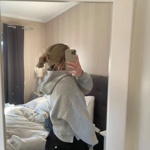 Grå croppad hoodie med huva - Säljer en grå croppad hoodie med huva och loose passform. Tröjan har långa ärmar och är tillverkad i mjukt bomullsmaterial, perfekt för chill dagar. Enkel och stilren design som funkar till jeans eller mjukisbyxor.