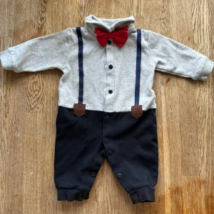 Grå och svart byxdress med rosett - Supersöt byxdress för baby med grå överdel, svarta byxben och en röd rosett vid halsen. Tryckknappar framtill och längs benen gör den enkel att ta på. Dekorativa mörkblå hängslen och bruna detaljer framtill. Mjuk bomullskänsla, perfekt för chill dagar.