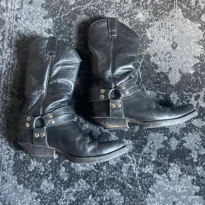 Svarta cowboy boots från Sendra - Riktigt snygga svarta cowboy boots från Sendra. Klassisk westernstil med dekorativa nitar och remmar runt ankeln. Boots med spetsig tå och låg klack, perfekta för dig som vill sticka ut med en cool look.