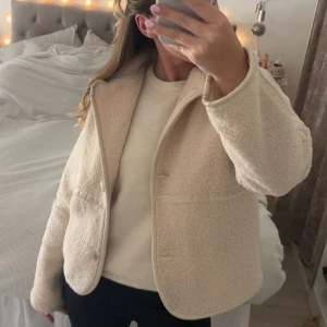 Beige teddyjacka med knappar - Supermysig beige teddyjacka med stora knappar framtill och markerade sömmar. Jackan har en boxig passform, lång ärm och två fickor framtill. Perfekt för lager-på-lager och ger en chill vibe till din outfit.
