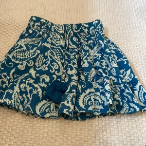 Blåvit kortkjol med tofsar - Supersöt kortkjol i blått och vitt med ett unikt blommigt mönster. Kjolen har resår i midjan och dekorativa tofsar framtill. Perfekt för varma dagar och ger en avslappnad men trendig vibe. Lätt och  passar perfekt till sneakers eller sandaler. Använt fåtal gånger