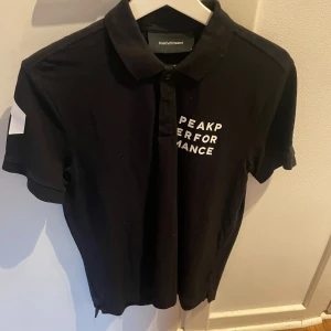 Svart Peak Performance t-shirt med tryck - Svart t-shirt från Peak Performance med vit texttryck på bröstet och en vit detalj på ärmen. Klassisk krage och knappslå framtill. Skön passform och stilren design, perfekt för dig som gillar sportiga och cleana plagg.