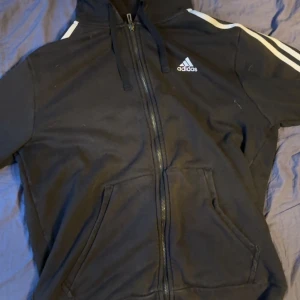 Svart Adidas hoodie med dragkedja - Svart hoodie från Adidas med klassiska vita ränder på axlarna och logga på bröstet. Tröjan har huva med snörning, hel dragkedja framtill och två fickor. Perfekt för chill eller träning, riktigt skön och sportig stil.