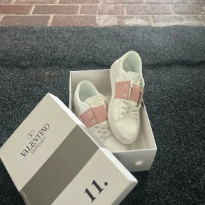 Valentino Garavani Open sneakers vit/rosa - Snygga Valentino Garavani Open sneakers i vitt skinn med bred rosa läderdetalj över snörningen. Klassisk rund tå, platt sula och ikoniska nitar baktill. Perfekta för dig som vill ha en lyxig och trendig look.
