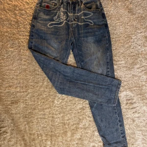 Billionaire Boys Club Jeans - Unika blå jeans med coolt tryck av en tecknad figur framtill och broderade detaljer på bakfickorna. Snygg röd brodyr på myntfickan. Jeansen har en rak passform och är tillverkade i klassiskt denimtyg. Perfekta för dig som vill sticka ut med din stil.