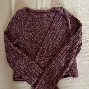 Vinröd omlottopp med ribbad struktur - Vinröd långärmad omlottopp med ribbad struktur och v-ringning. Toppens material är mjukt och stretchigt, perfekt för en chill och snygg look. Croppad modell som passar till både jeans och kjol. Ingen synlig logga eller märke.