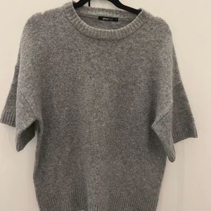 Grå stickad tröja från Gina Tricot - Mysig grå stickad tröja från Gina Tricot med korta ärmar och rund halsringning. Tröjan har en loose passform och breda ribbade muddar vid ärmslut och nederkant. Perfekt för lager-på-lager och chill dagar.