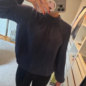 Mörkblå blus med hög krage - Säljer en mörkblå blus med lång ärm och snygg hög krage. Blusen har lätt glansigt tyg.