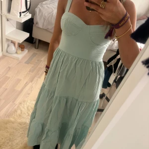 Ljusblå långklänning med smockrygg - Supersöt ljusblå långklänning med breda axelband och markerad byst. Klänningen har smock i ryggen för extra komfort och volangig kjol i flera lager. Perfekt för varma dagar och har en luftig känsla. Materialet är lätt och känns som bomull.