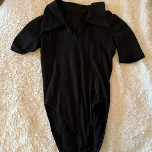 Svart body med krage och v-ringning - Svart body med korta ärmar, v-ringning och stor krage. Snygg och enkel design som passar till mycket. Knäppning nedtill för enkel på- och avtagning. Perfekt att styla med jeans eller kjol.