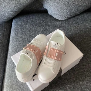 Valentino Rockstud sneakers vit/rosa - Snygga vita Valentino Rockstud sneakers med breda ljusrosa band och ikoniska nitar över vristen. Skorna har rund tå, platt sula och är gjorda i skinn. Perfekta för dig som vill sticka ut med en lyxig och edgy vibe.