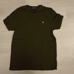 Olivgrön t-shirt från Lyle & Scott - Snygg olivgrön t-shirt från Lyle & Scott med klassisk gul logga på bröstet. Modellen är rak med rund hals och korta ärmar. Mjuk och skön bomull som passar perfekt till jeans eller shorts. Enkelt och stilrent plagg med cool vibe.