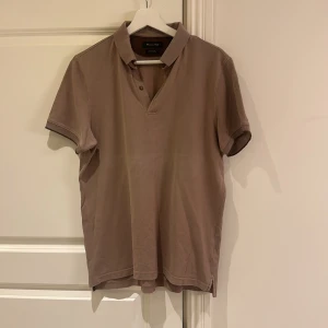 Beige pikétröja från Massimo Dutti - Snygg beige pikétröja från Massimo Dutti med klassisk krage och två knappar vid halsen. T-shirten har korta ärmar och är gjord i mjuk bomull som känns skön mot huden. Perfekt för en chill och stilren look.