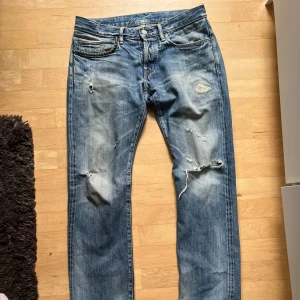 Blå distressed jeans från Hysteric Glamour - Säljer ett par blå jeans från Hysteric Glamour med slitna detaljer och hål på benen för en riktigt edgy look. Klassisk femficksmodell med läderpatch bak i midjan. Jeansen har en rak passform och är tillverkade i robust denim.