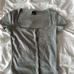 Grå ribbad topp från Gina Tricot - Grå ribbad topp från Gina Tricot med korta ärmar och knappar framtill. Toppen har en fyrkantig urringning och är figurnära i modellen. Perfekt att styla med jeans eller kjol för en enkel och snygg look. Inte änvänd många gånger, jätte bra skick alla knappar funkar!💕