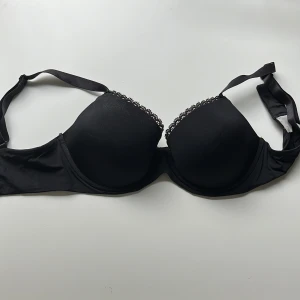 Svart axelbandslös bandeau-bh - Snygg svart bandeau-bh med vadderade kupor och avtagbara axelband. Ovanför kuporna finns en fin spetskant som ger en extra touch. Perfekt under toppar med bara axlar eller offshoulder-plagg.