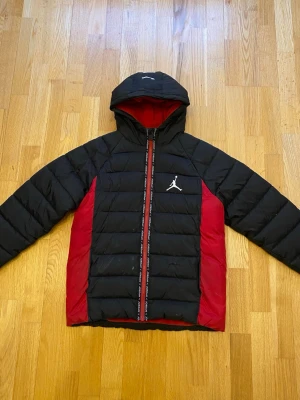 Jordan svart och röd pufferjacka  - Säljer en fet svart och röd pufferjacka från Jordan med huva och ikoniska Jumpman-loggan på bröstet. Jackan har quiltad design, dragkedja framtill och röda paneler på sidorna. Perfekt för kalla dagar och riktigt snygg streetstil.