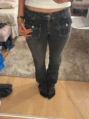 Svarta jeans med fickdetaljer - Säljer ett par svarta jeans med snygga fickor framtill med lock och knapp. Jeansen har raka ben och klassisk femficksdesign baktill. De är lowwaist.