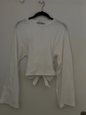 Vit croppad topp med knyt i ryggen - Säljer en vit croppad topp med långa ärmar och snygg knytning i ryggen som ger en unik look. Toppen har en enkel, stilren design och är gjord i mjukt bomullstyg. Perfekt för dig som vill ha något trendigt och bekvämt. Har endast använt en gång i skolan