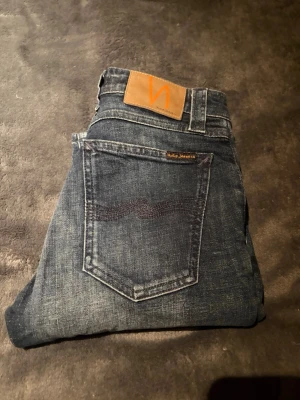  Nudie jeans  - Säljer ett par mörkblå skinny jeans från Nudie med klassisk femficksdesign och snygga slitningar. Jeansen har kontrastsömmar, läderpatch med logga bak och är tillverkade i mjukt jeansmaterial. Perfekta för dig som gillar en smal passform och stilren look.