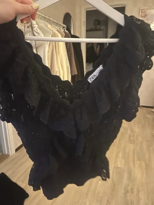 Svart spetsblus med volang från Zara - Säljer en svart blus från Zara i spets med stora volangdetaljer längs ringningen. Blusen är ärmlös och har en romantisk look med genombrutet mönster. Perfekt för dig som gillar detaljer och vill sticka ut lite extra.