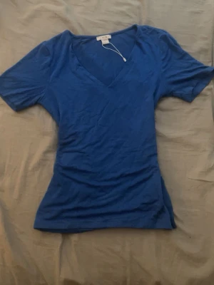 Blå v-ringad topp  - En enkel och snygg blå topp med v-ringning och korta ärmar. Modellen är figurnära och passar perfekt till jeans eller kjol. T-shirten är gjord i mjukt material som känns skönt mot huden.