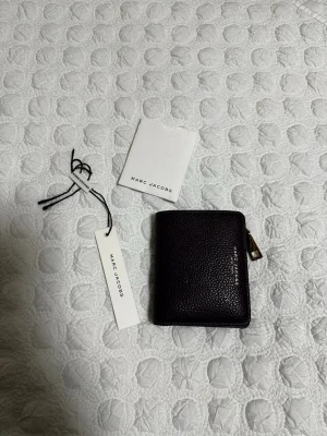 Mörklila plånbok från Marc Jacobs - Snygg mörklila plånbok från Marc Jacobs i äkta skinn. Plånboken har flera kortfack, ett myntfack med dragkedja i guld och tryckknappsstängning. Stilren design med Marc Jacobs-logga på framsidan. Perfekt storlek för fickan eller väskan.