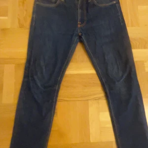 Mörkblå jeans från Nudie Jeans W32 L32 - Säljer ett par mörkblå jeans från Nudie Jeans i klassisk femficksmodell. Jeansen har raka ben och orangea sömmar som ger en snygg kontrast. Tillverkade i slitstarkt bomullstyg och passar perfekt till en avslappnad stil.