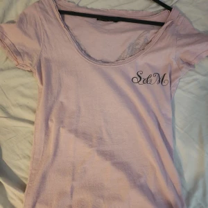 Ljusrosa t-shirt Saints & Mortals - Ljusrosa t-shirt från Saints & Mortals med coolt svart tryck på ryggen med döskalle, blommor och texten 'Sinners'. Framsidan har diskret logga vid bröstet. T-shirten har korta ärmar och är i mjuk bomull, perfekt för en edgy streetstil. Passar S/M