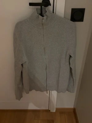 Ljusgrå kofta från Selected Homme - Ljusgrå stickad kofta från Selected Homme med hög krage och hel dragkedja framtill. Tröjan har ribbade muddar och en clean, minimalistisk look som passar perfekt till lager på lager under höst och vinter.