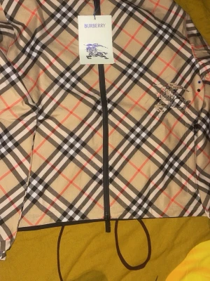 Rutig Burberry vindjacka med huva - Säljer en snygg vindjacka från Burberry med klassiskt rutig mönster i beige, svart, vit och röd. Jackan har huva, dragkedja framtill och Burberry-logga broderad på bröstet. Perfekt för dig som vill sticka ut med stilren design och ikoniskt mönster.