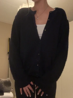 Mörkblå mysig kofta med knappar - Mysig mörkblå kofta med rund hals och knäppning framtill. Koftan har en loose passform och är gjord i ett fluffigt stickat material som känns riktigt skönt. Perfekt att slänga på sig över ett linne eller t-shirt. Långa ärmar och enkel, clean look.