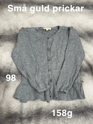 Grå kofta med guldprickar, stl 98 - Supersöt grå kofta med små guldprickar och volangkant nedtill. Koftan har knappar framtill och är långärmad, perfekt för att piffa upp vardagslooken. Mjuk och skön stickad känsla som passar till allt.🌸 Fint använt skick. Guldiga prickar. Storlek 98