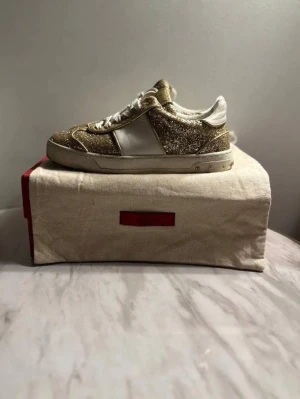Valentino glitter sneakers guld/vit - Säljer ett par Valentino sneakers med glittriga guldfärgade paneler och vita detaljer. Skorna har snörning, platt sula och klassisk rund tå. Märkets ikoniska nitar syns på sulan. Perfekta för dig som vill sticka ut med exklusiv design och coola detaljer. En nit saknas. Pris diskuterbart 