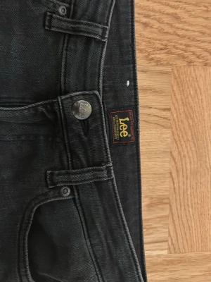Svarta Lee Jessica bootcut jeans W25 L31 - Säljer ett par svarta Lee Jessica jeans med bootcut passform. Klassisk femficksmodell med Lee-logga på bakfickan och knapp. Jeansen är i stretchig bomullsdenim och har en snygg mörk tvätt. Perfekta för dig som gillar en tidlös look med lite utsvängda ben.