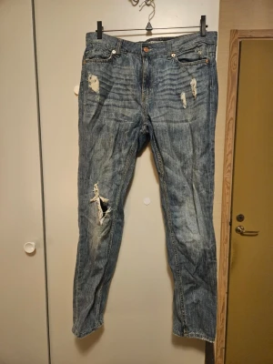 Blå slitna jeans från Lindex - Säljer ett par blå jeans från Lindex med slitningar och hål på både fram- och baksida. Klassisk femficksmodell med normal passform och raka ben. Jeansen har en snygg tvättad look och detaljerade slitningar för en avslappnad stil.