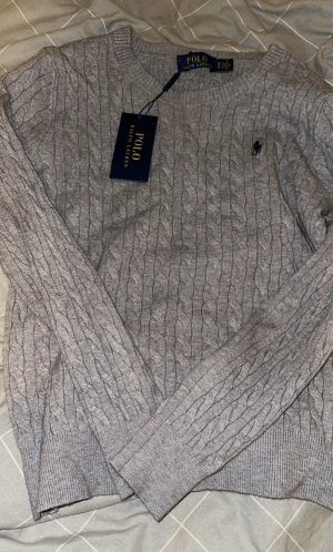 Grå kabelstickad tröja Polo Ralph Lauren - Barn storlek s mindre i storlek Säljer en grå kabelstickad tröja från Polo Ralph Lauren med klassisk rund hals och diskret logga på bröstet. Tröjan har långa ärmar och ribbade muddar. Perfekt för dig som gillar stilrena och tidlösa plagg.