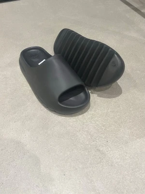 Svarta Adidas slides i syntet - Säljer ett par svarta Adidas slides med öppen tå och chunky sula. Tillverkade i syntetmaterial och har en clean, minimalistisk look med Adidas-logga på innersulan. Perfekta för chill dagar eller efter träningen.