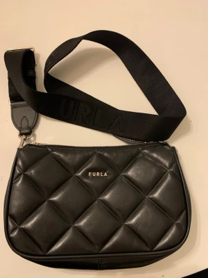 Svart quiltad axelväska från Furla - Säljer en svart axelväska från Furla i äkta skinn med quiltad design och bred justerbar axelrem med Furla-logga. Väskan har dragkedja upptill och en stilren look som passar perfekt till din streetstyle. Perfekt storlek för det viktigaste.  En fin axelbandsväska från Flura. Knappt använd.   Mått:  Bredd: ca 23 cm  Längd: 15 cm  Vidd: 5 cm
