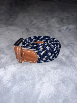Flätat blått och vitt bälte - Säljer ett snyggt flätat bälte i marinblått, vitt och grått med detaljer i brunt konstläder och silverfärgat spänne. Perfekt accessoar för att lyfta din outfit med en sportig och stilren touch.